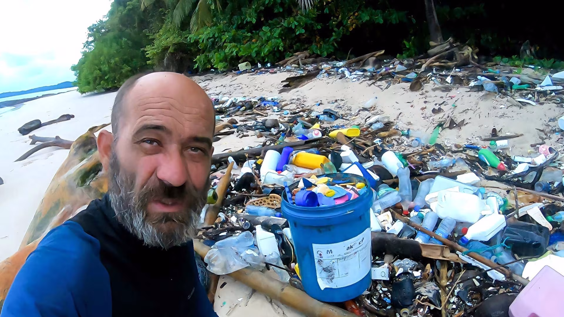 Un homme debout, sur une plage face à l'océan devant un grand tas de déchets.